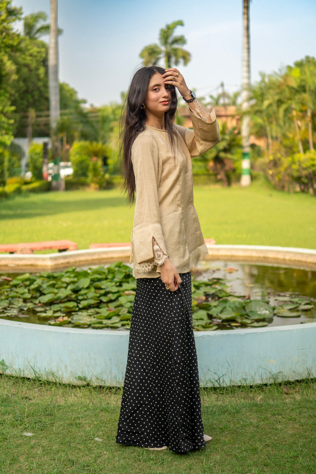 Woman in beige top and black polka dot skirt standing in a garden with a pond and greenery.