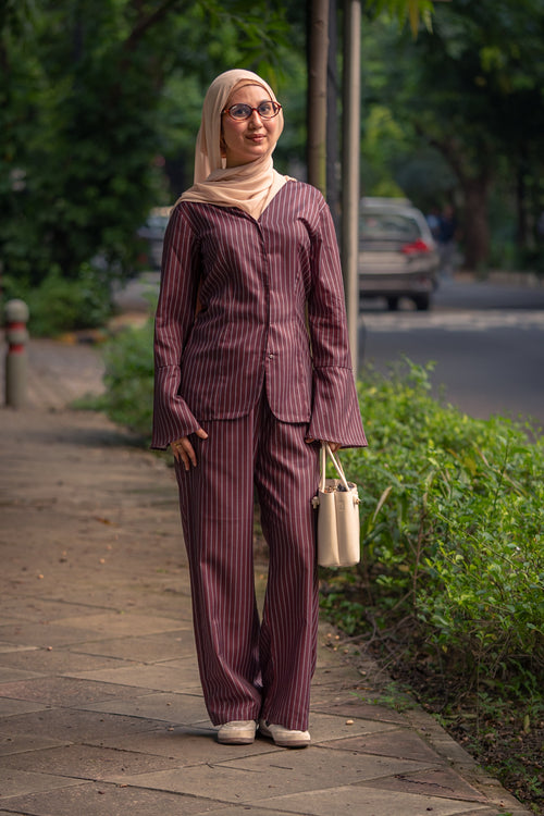 Woman in a maroon striped outfit walking on a sidewalk with greenery and a road in the background.